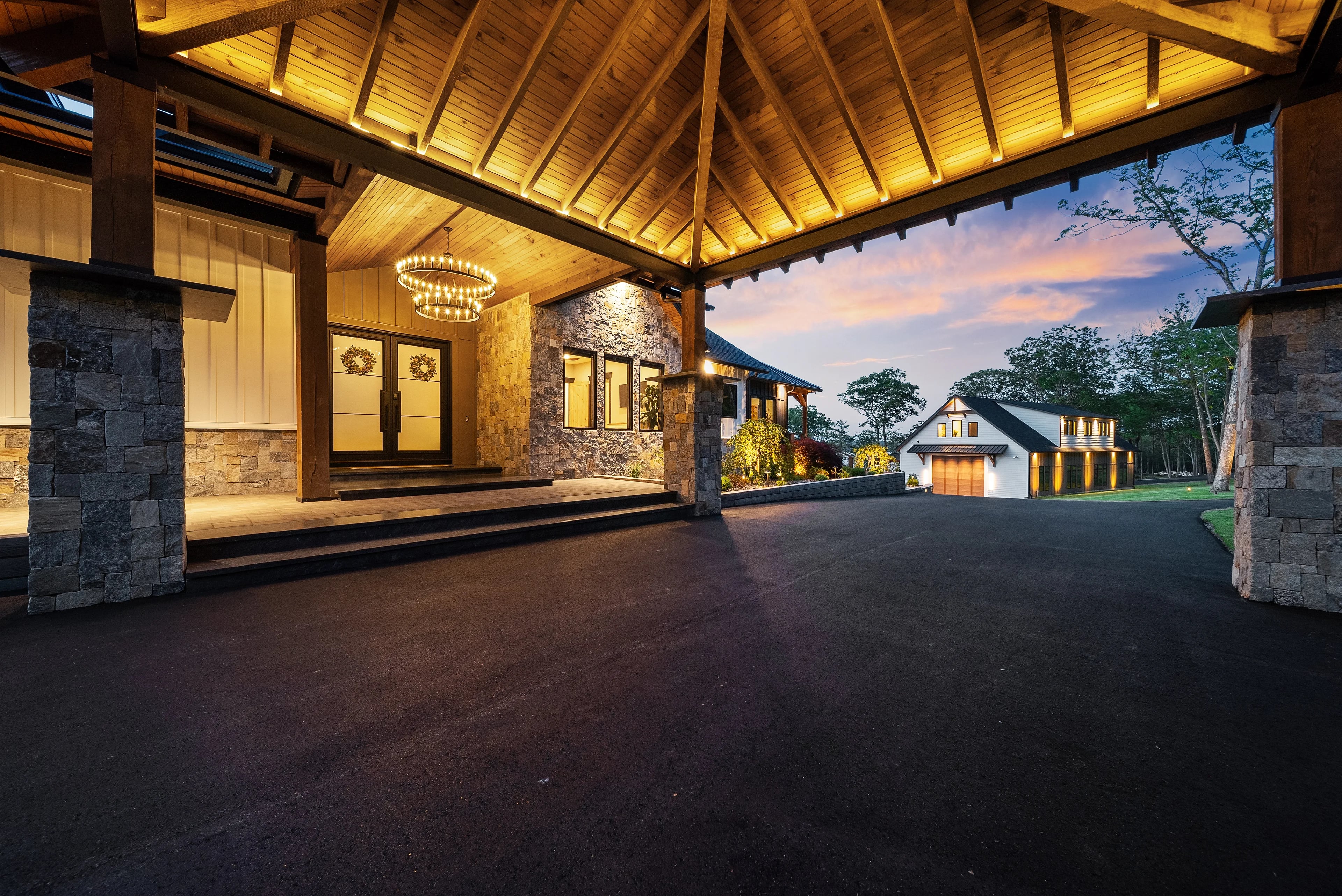 Under the porte-cochère at dusk, timber rafters radiating overhead between stone columns that frame the carriage house beyond