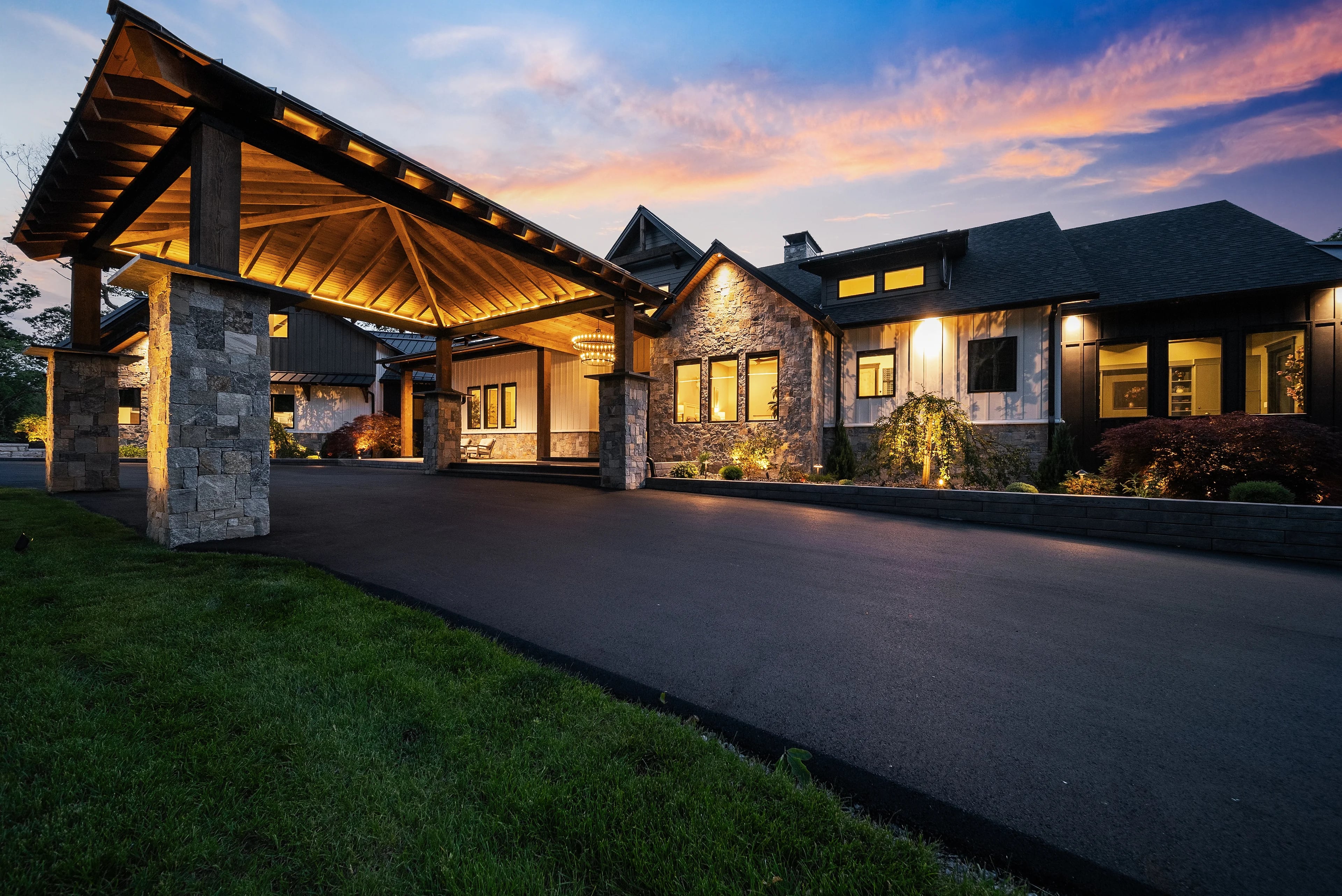 Twilight exterior of a timber-and-stone lakefront home, the lit porte-cochère drawing the eye down a sweeping drive under a sunset sky