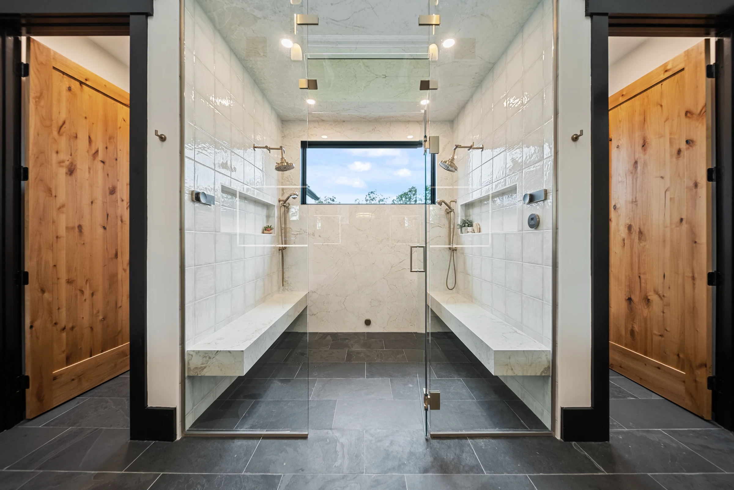 Mirrored dual shower in marble tile, matching benches and fixtures either side of a centred window, alder doors flanking the opening