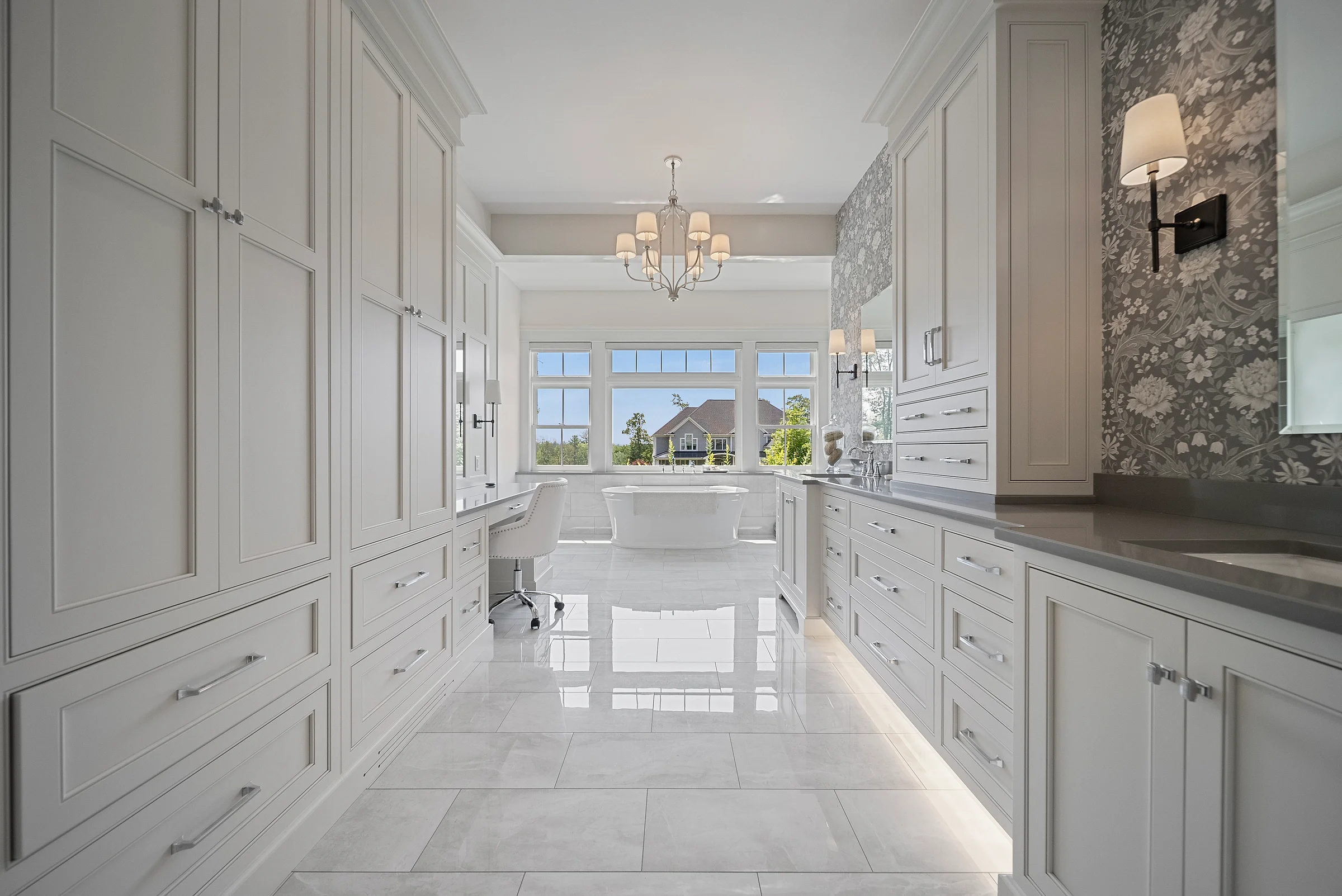 Fitted dressing corridor in pale cabinetry running to a bright bath beyond, a chandelier centred over the passage and stone tile underfoot