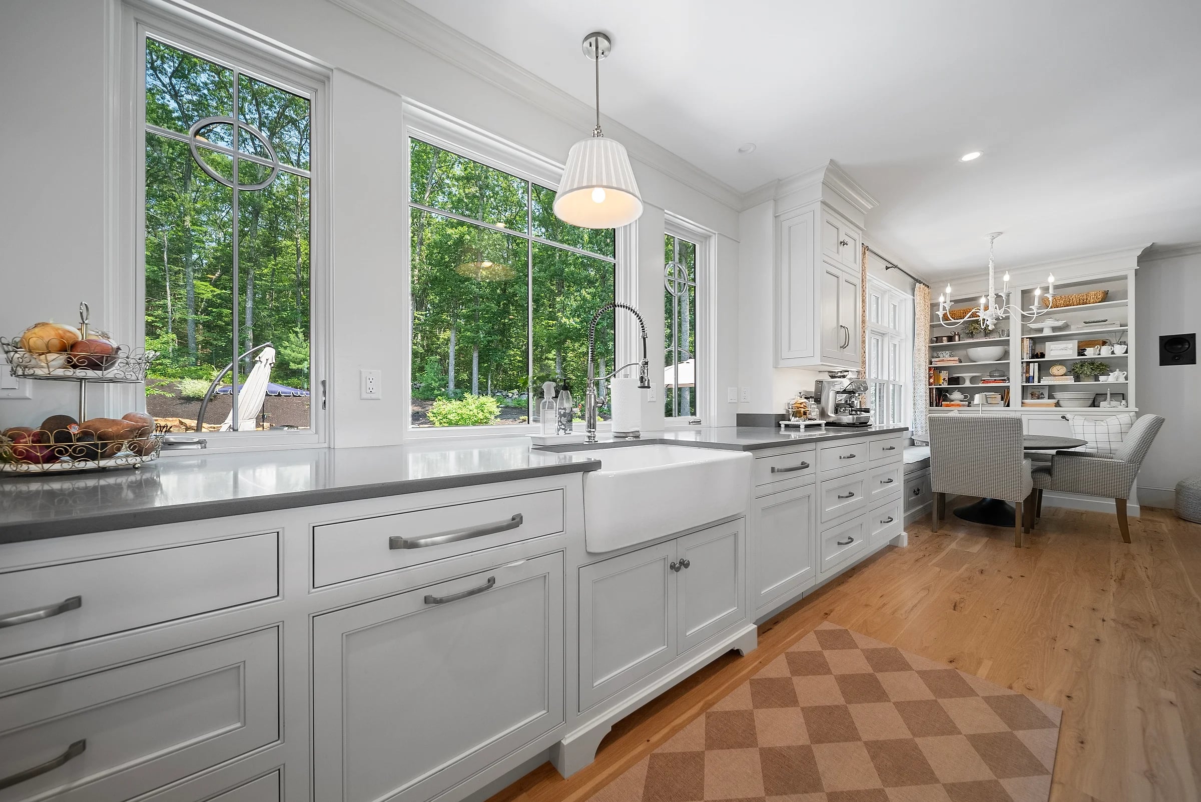 White inset-panel kitchen under a run of tall windows, a farmhouse sink and honed counters facing the treeline, checkerboard inlay set into the wood floor