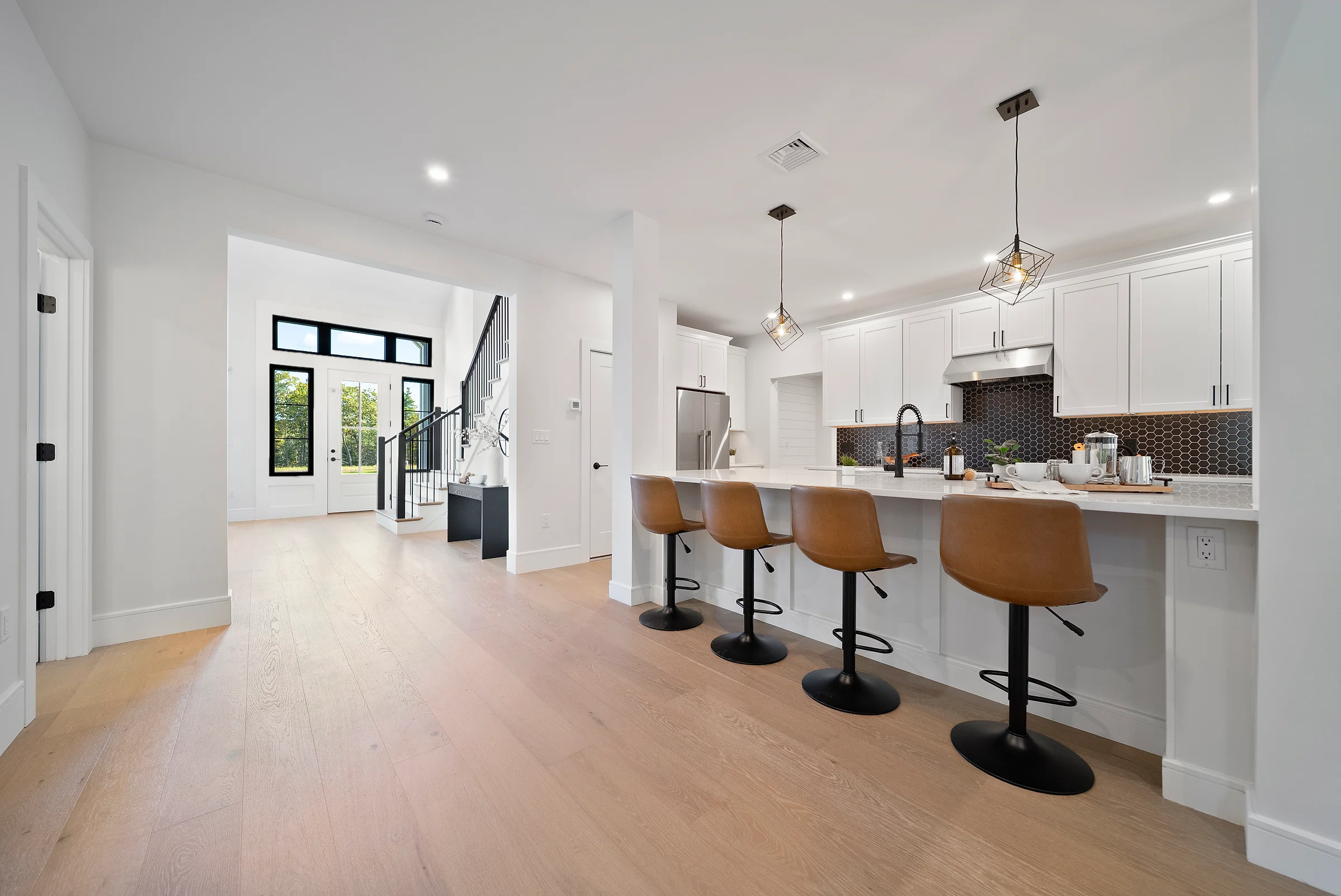 Open-plan kitchen in white shaker cabinetry with a long island and leather stools, wide-plank oak running back to a glazed entry