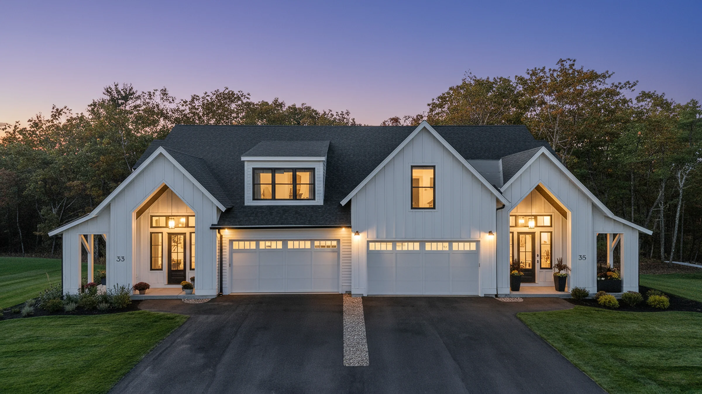 Paired white farmhouse elevation at twilight, gabled dormers and lit windows over a shared drive against a violet sky