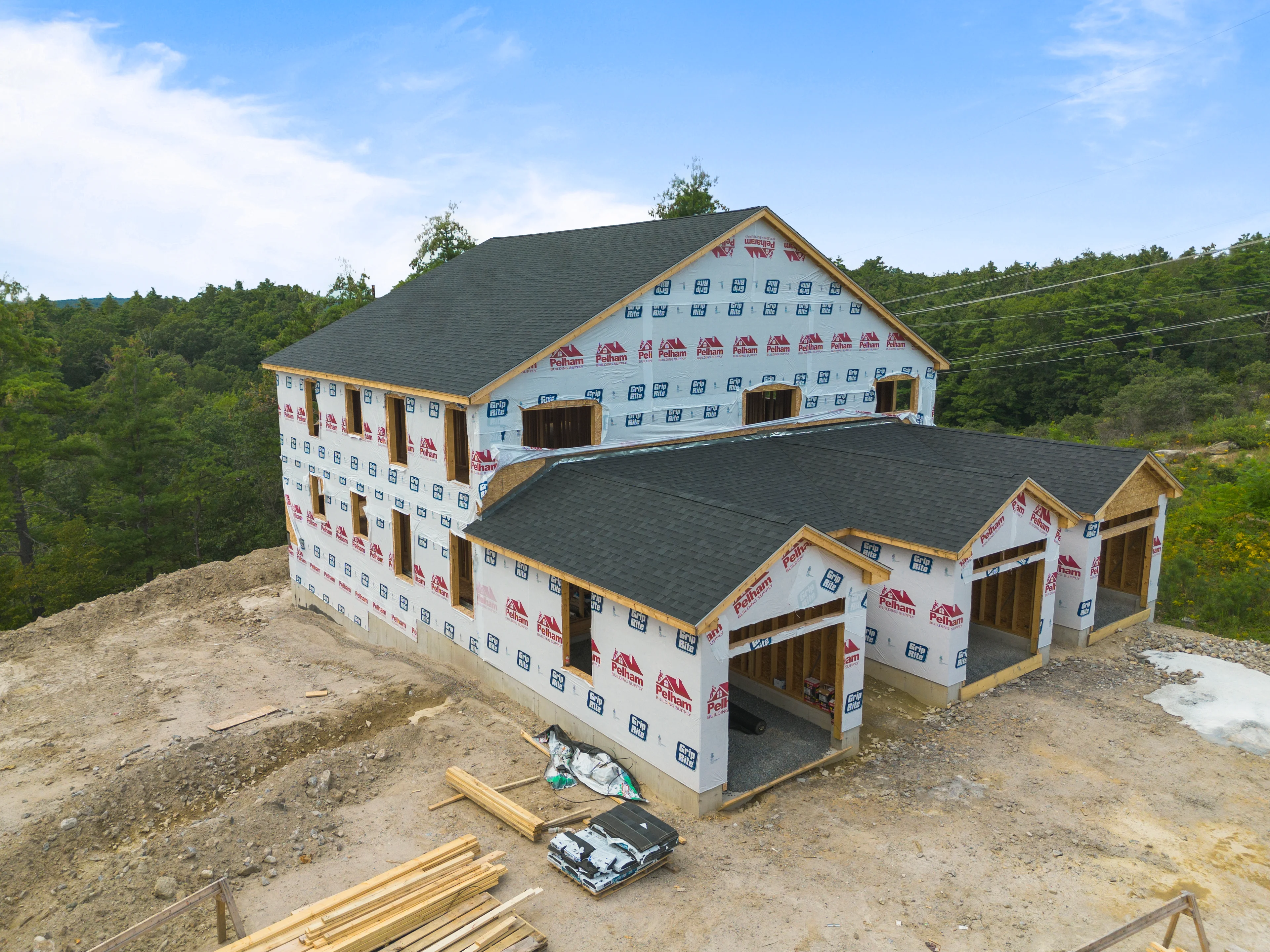 Aerial of a house fully wrapped and roofed with its garage bays still open framing, lumber and material bundles staged on the raw cut