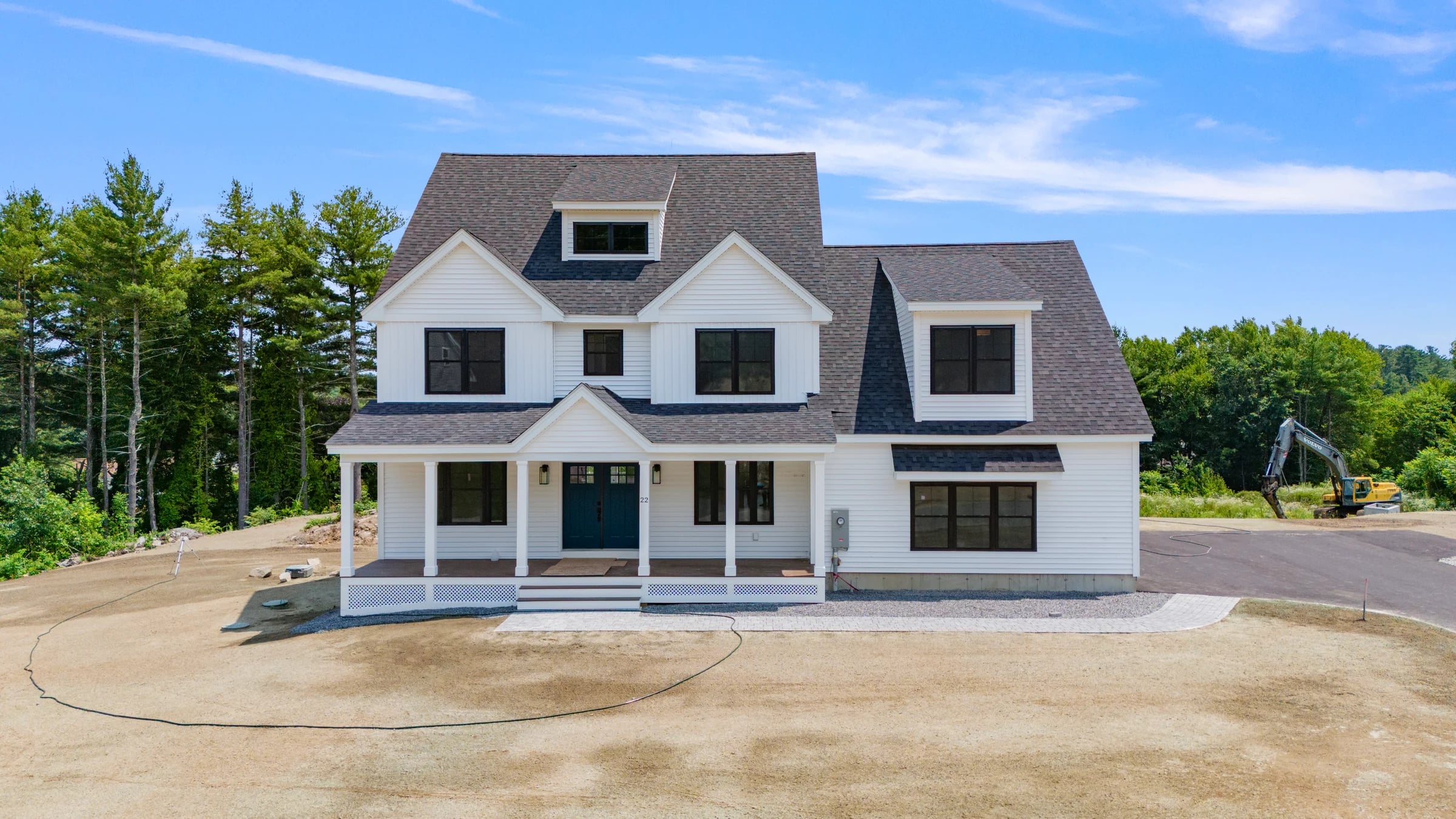 Finished white farmhouse elevation with the site still raw — graded dirt, grade stakes, and an excavator at the treeline