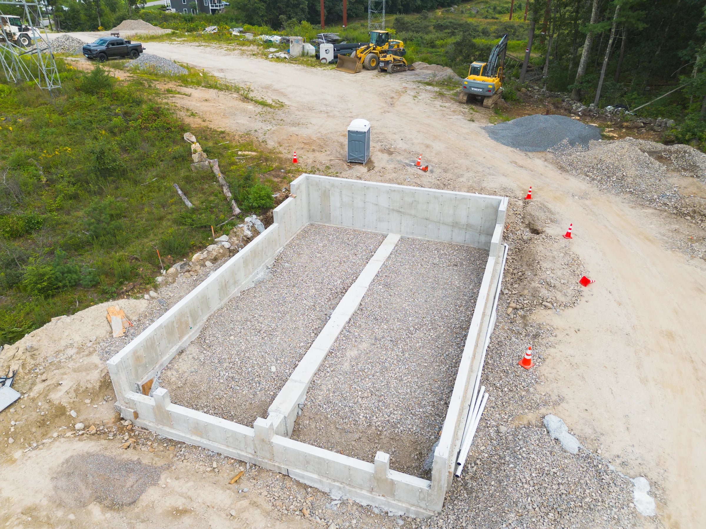 Overhead view of a poured concrete foundation with gravel fill and a centre footing, an excavator and wheel loader parked on the pad beyond