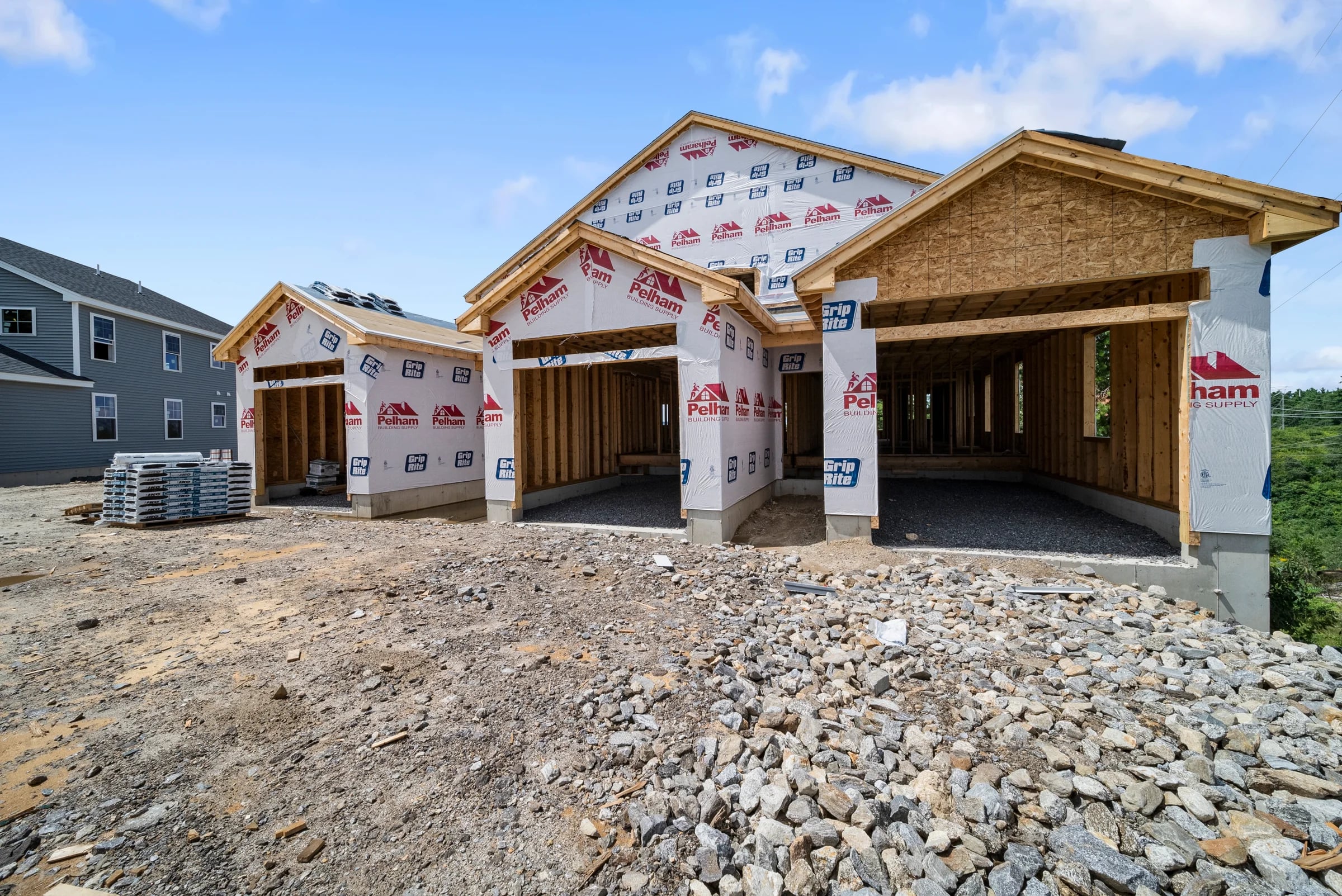 Ground view of a house in branded house-wrap with open framed garage bays, sheathed gable, and material pallets stacked on the gravel pad
