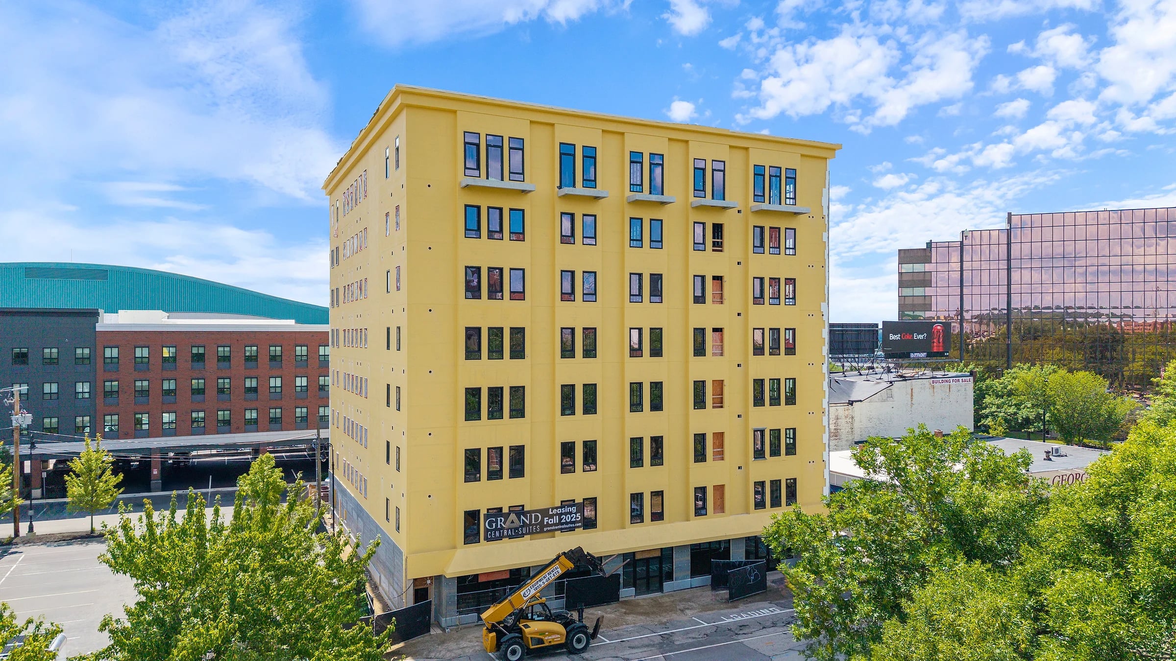 The finished seven-storey mid-rise in its new yellow facade with ground-floor retail signage, a telehandler still parked at the kerb