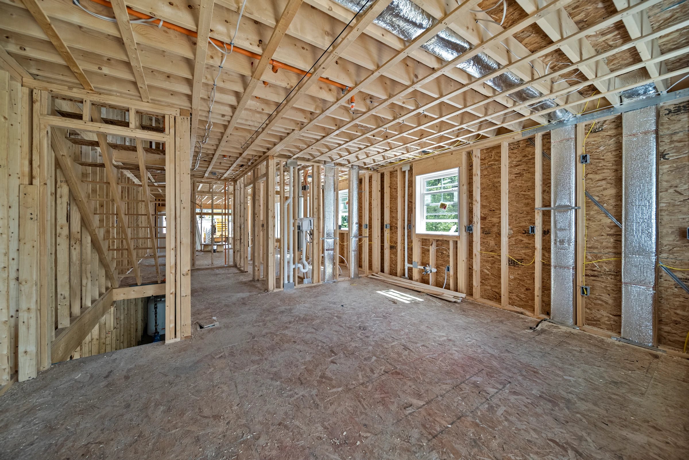 Interior at framing stage — open stud walls, floor joists overhead, ductwork, plumbing stacks, and electrical rough-in