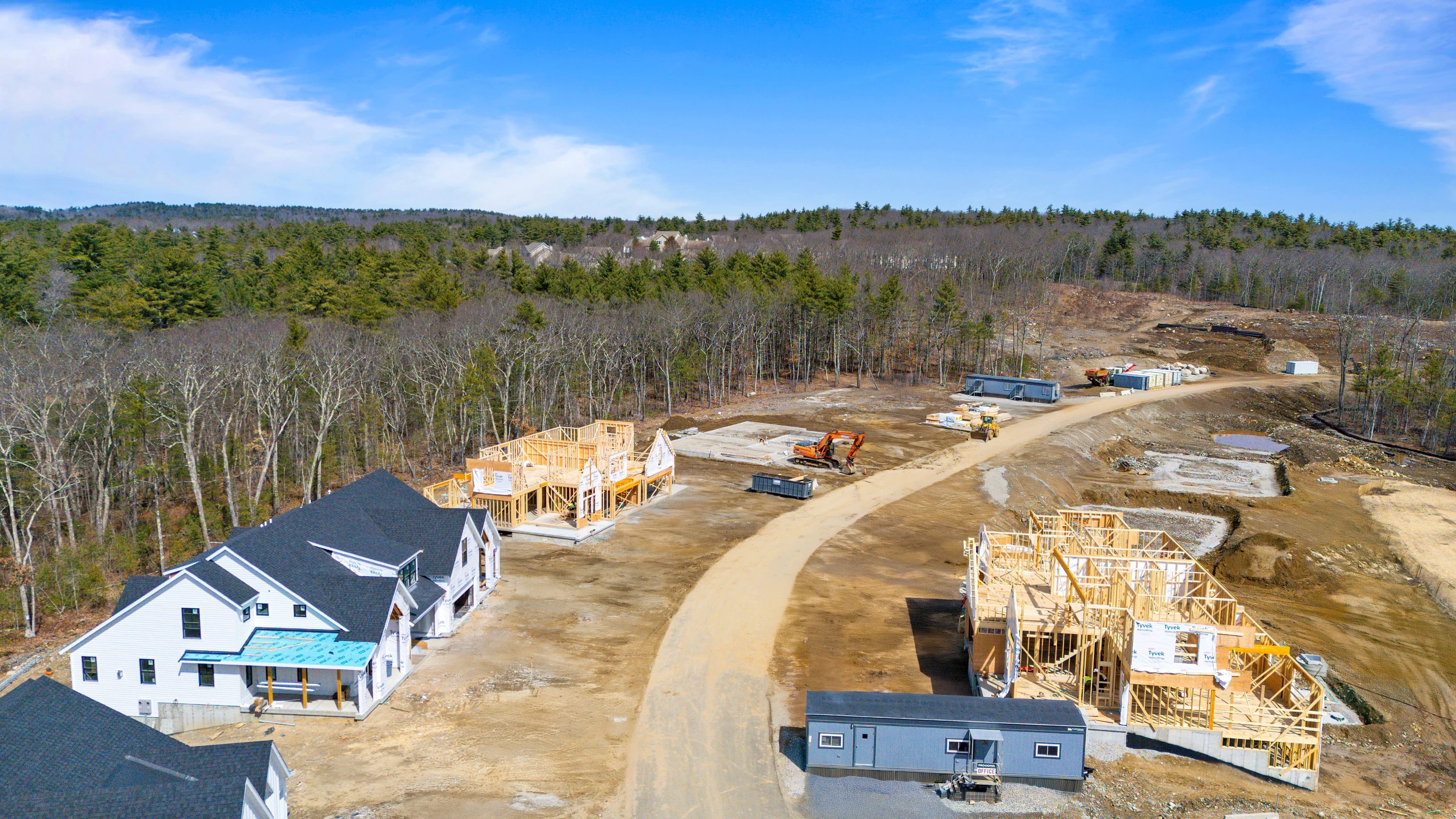 Aerial of an active subdivision with a finished home, two homes under framing, a poured slab, an excavator, and site trailers