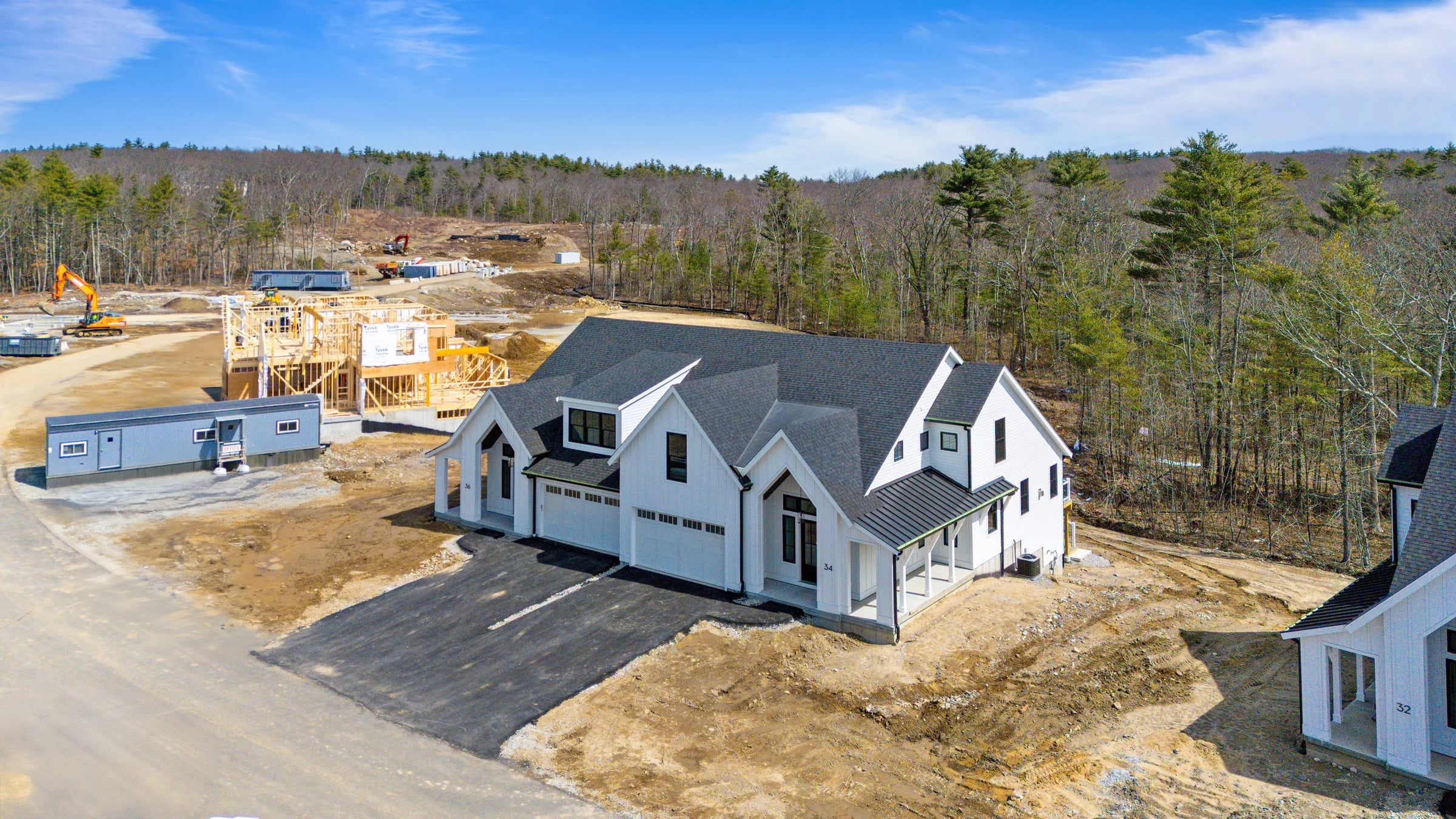 Aerial across a development with one home finished, another still open framing, an excavator working the graded dirt between them