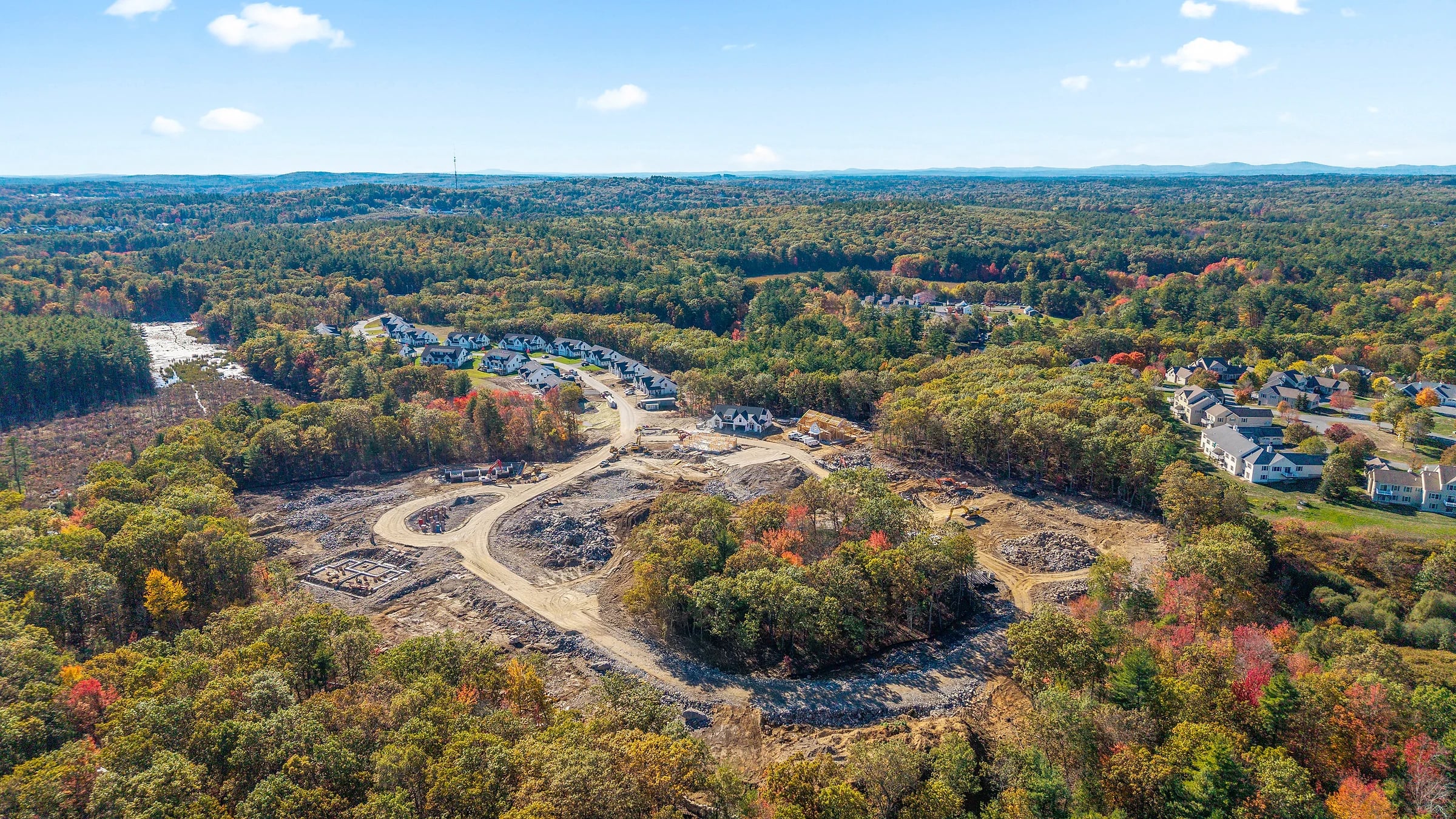 Aerial of a development site cleared and graded out of autumn forest, stockpiles and haul roads across the cut with an established neighbourhood beyond