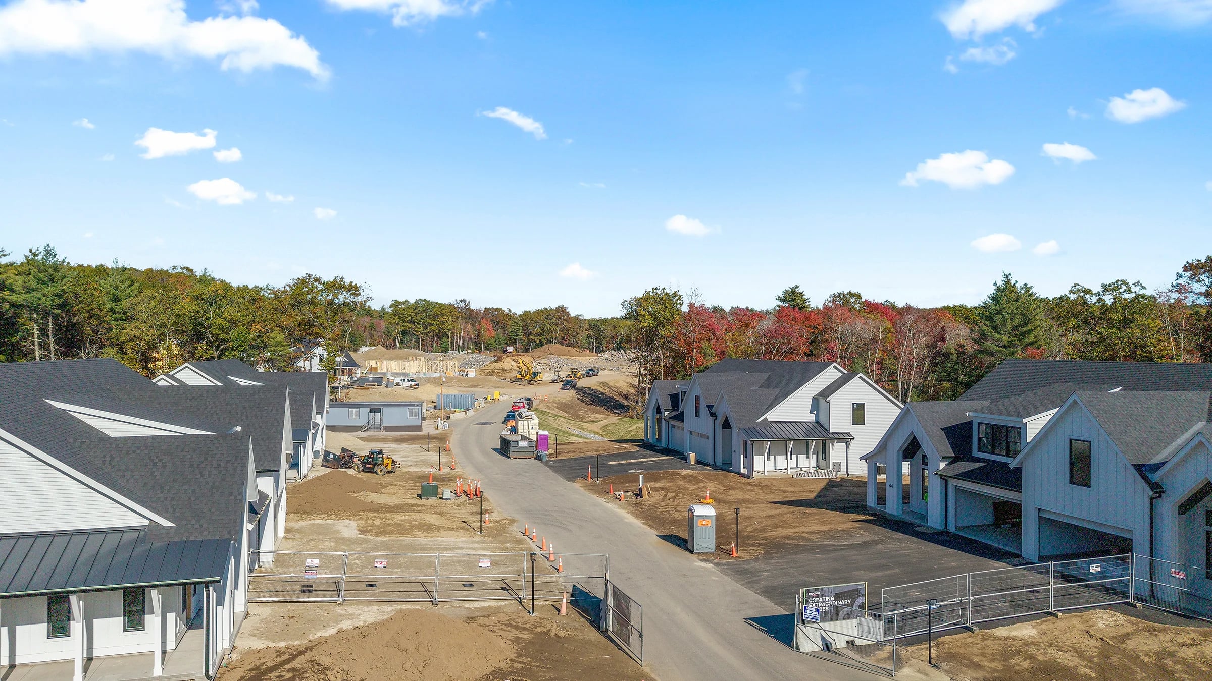 A new street of gabled homes under construction, graded roadway and raw dirt lots between them, autumn treeline beyond