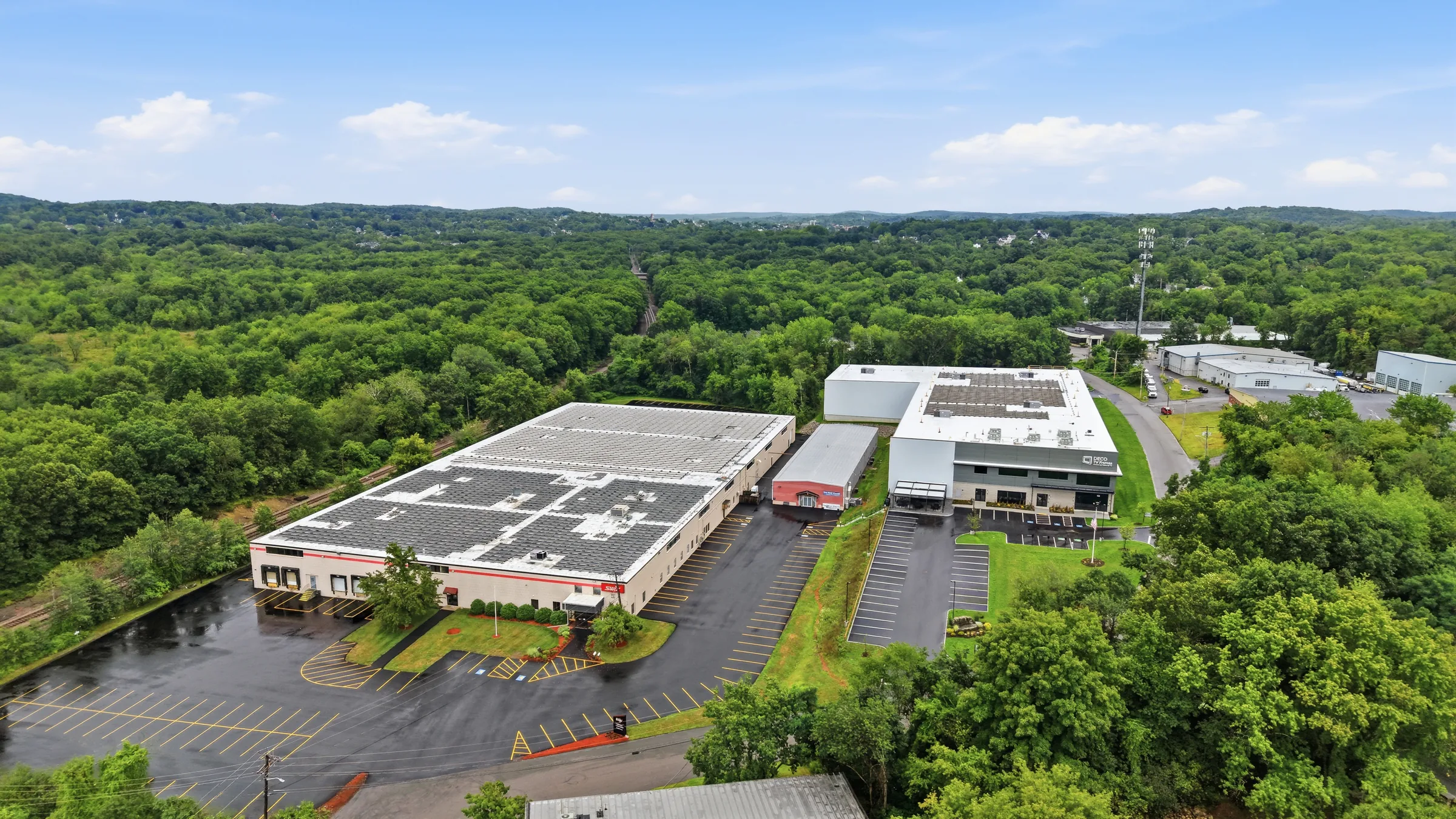 An aerial view of a manufacturing campus, two long buildings with parking, rail line, and woodland beyond