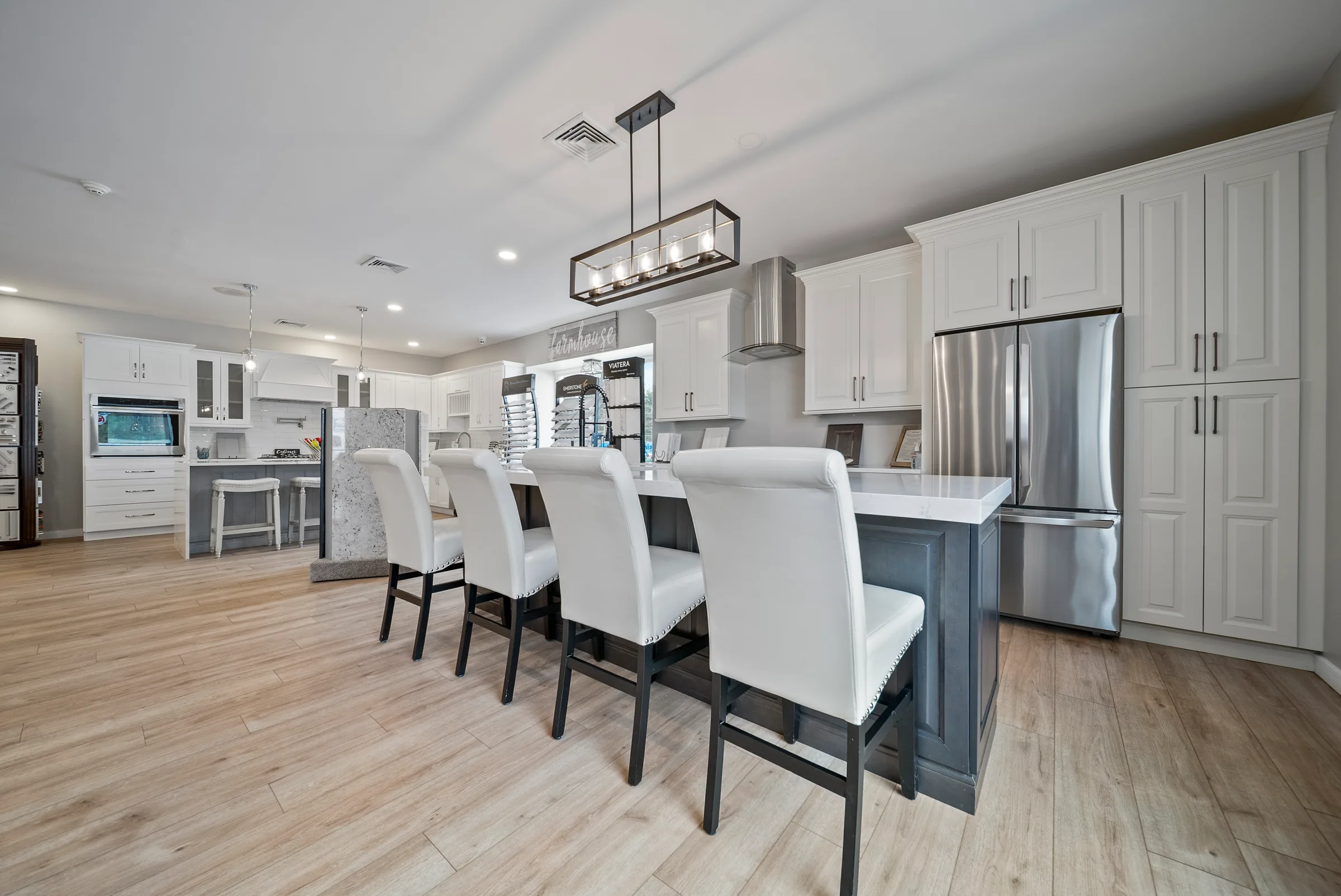 A showroom kitchen vignette with white shaker cabinets, a dark island and four upholstered barstools beneath a linear pendant