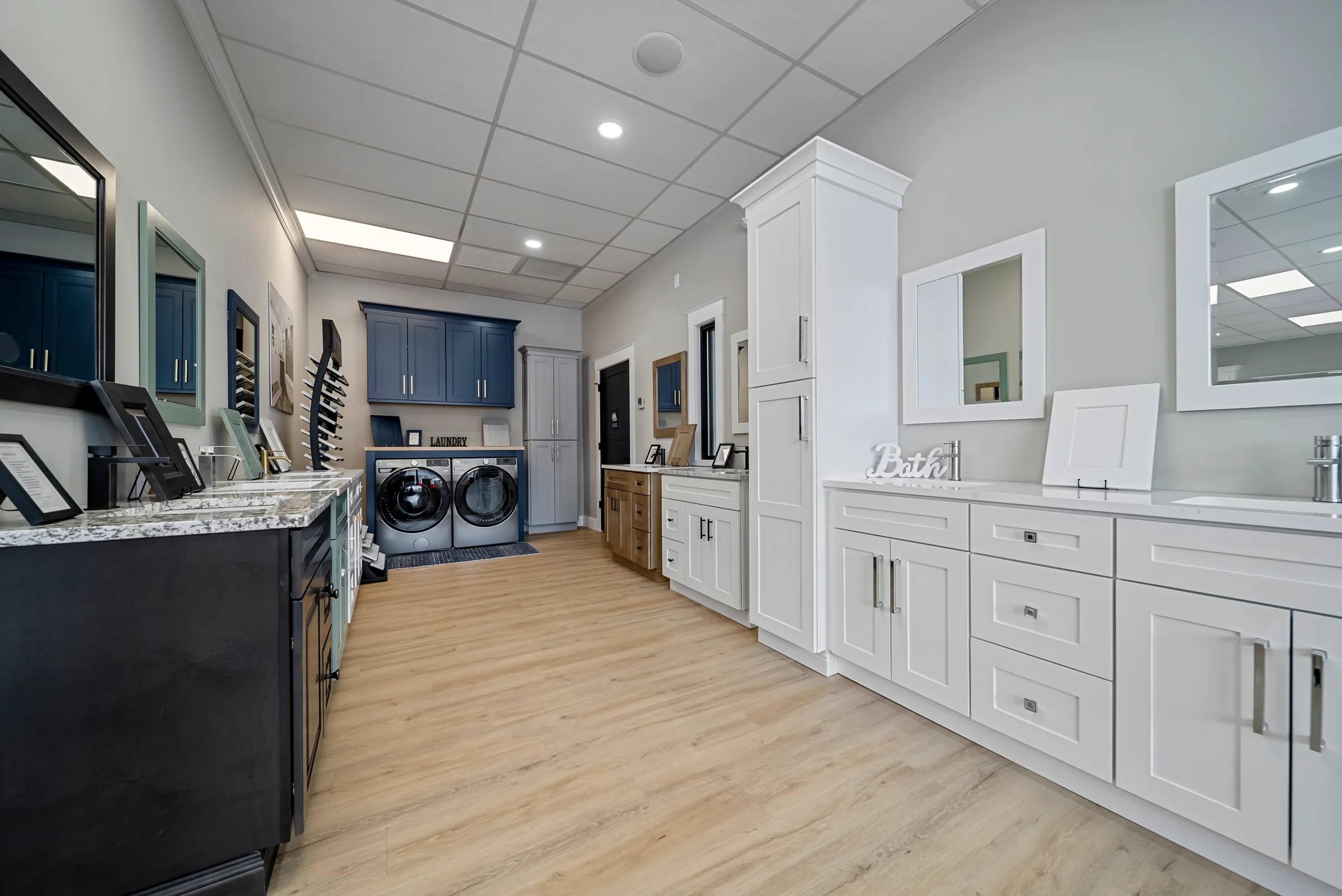 A showroom's bath and laundry display, white vanity cabinets and framed mirrors along one wall with a blue-grey laundry setup beyond