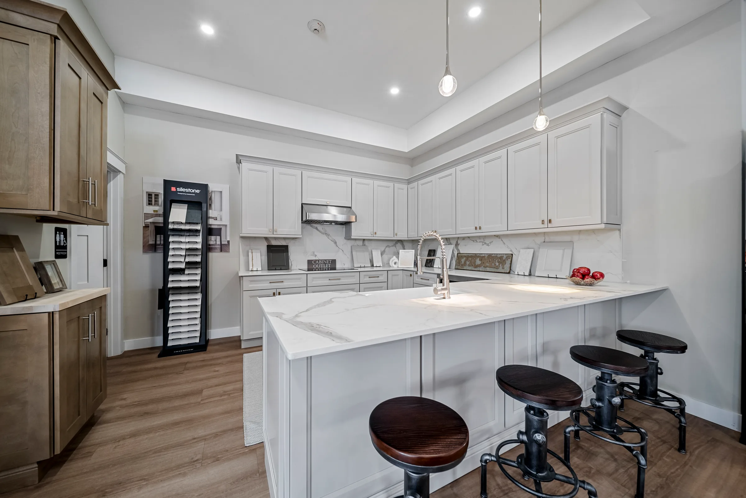 A showroom kitchen with a marble waterfall island, three metal stools and a door-sample rack against stained wood cabinetry
