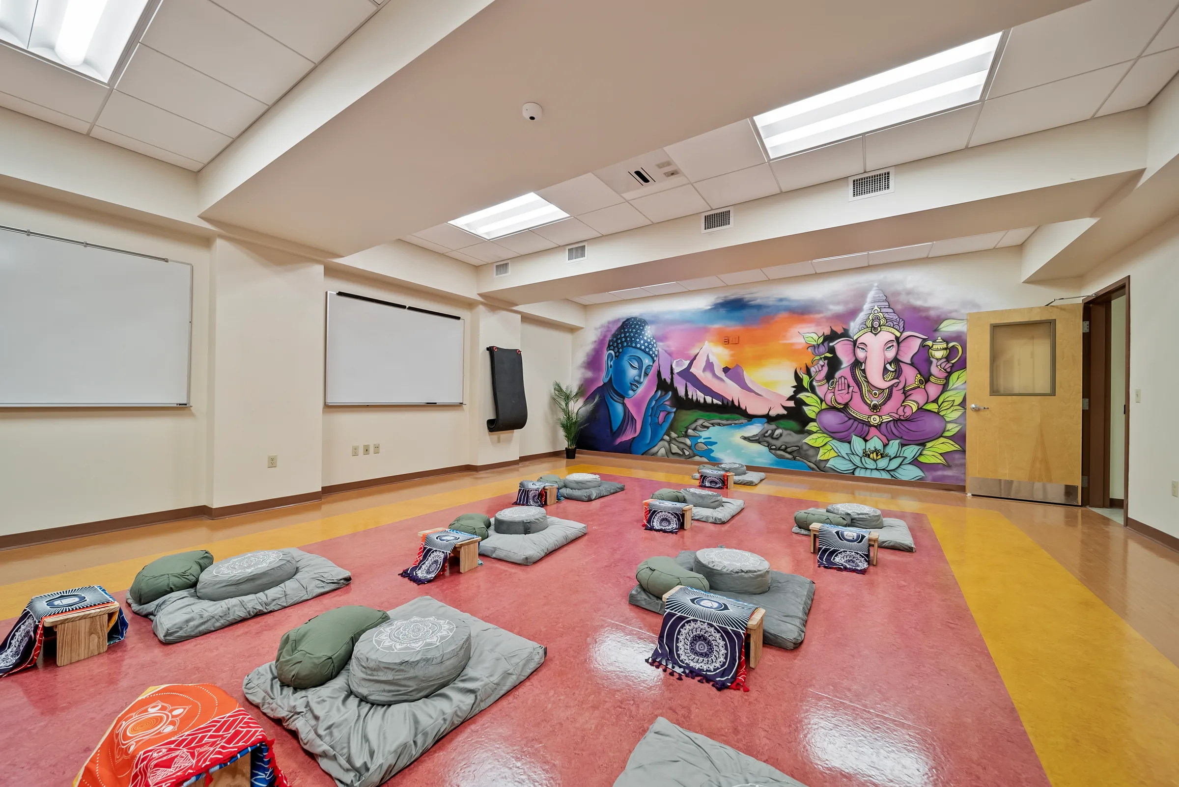 A group room with floor cushions arranged on a pink floor before a large painted mountain mural, a whiteboard on the adjacent wall