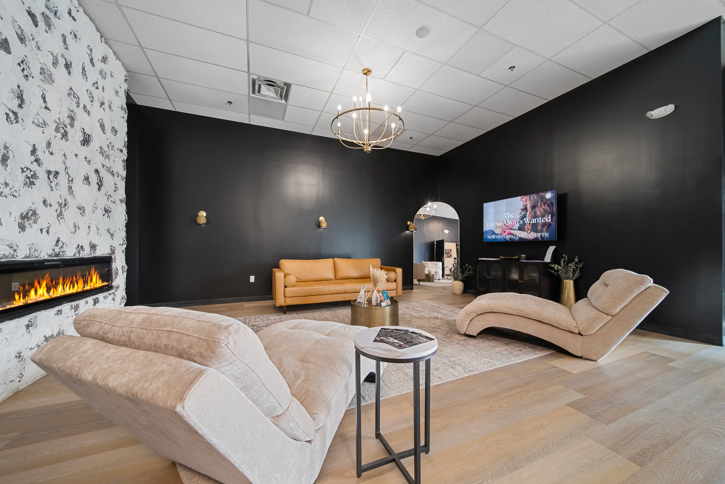A dark-walled lounge with a linear fireplace, chaise seating, and a chandelier