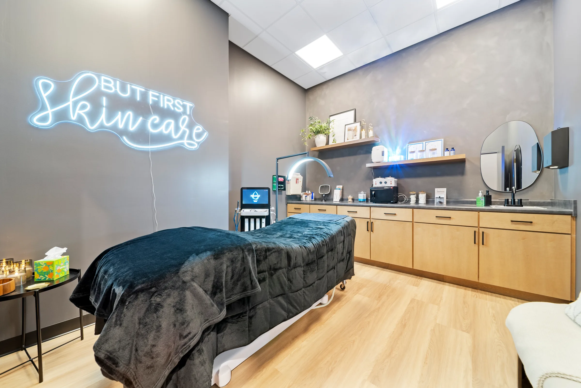 A treatment room with a draped bed and a neon sign reading But First Skincare above the counter