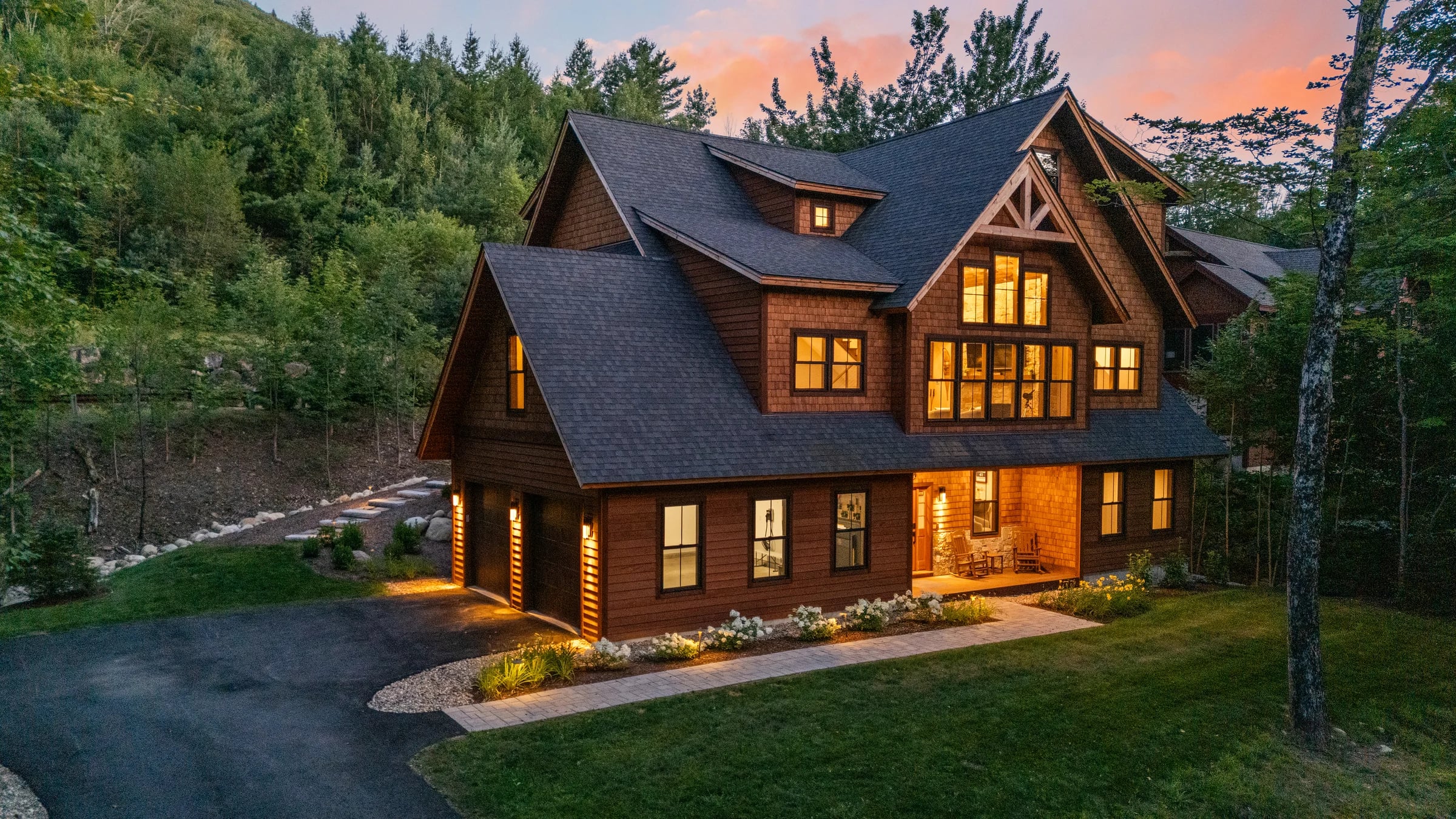 A dark cedar lodge lit warm against the twilight, its landscaped entry in the foreground