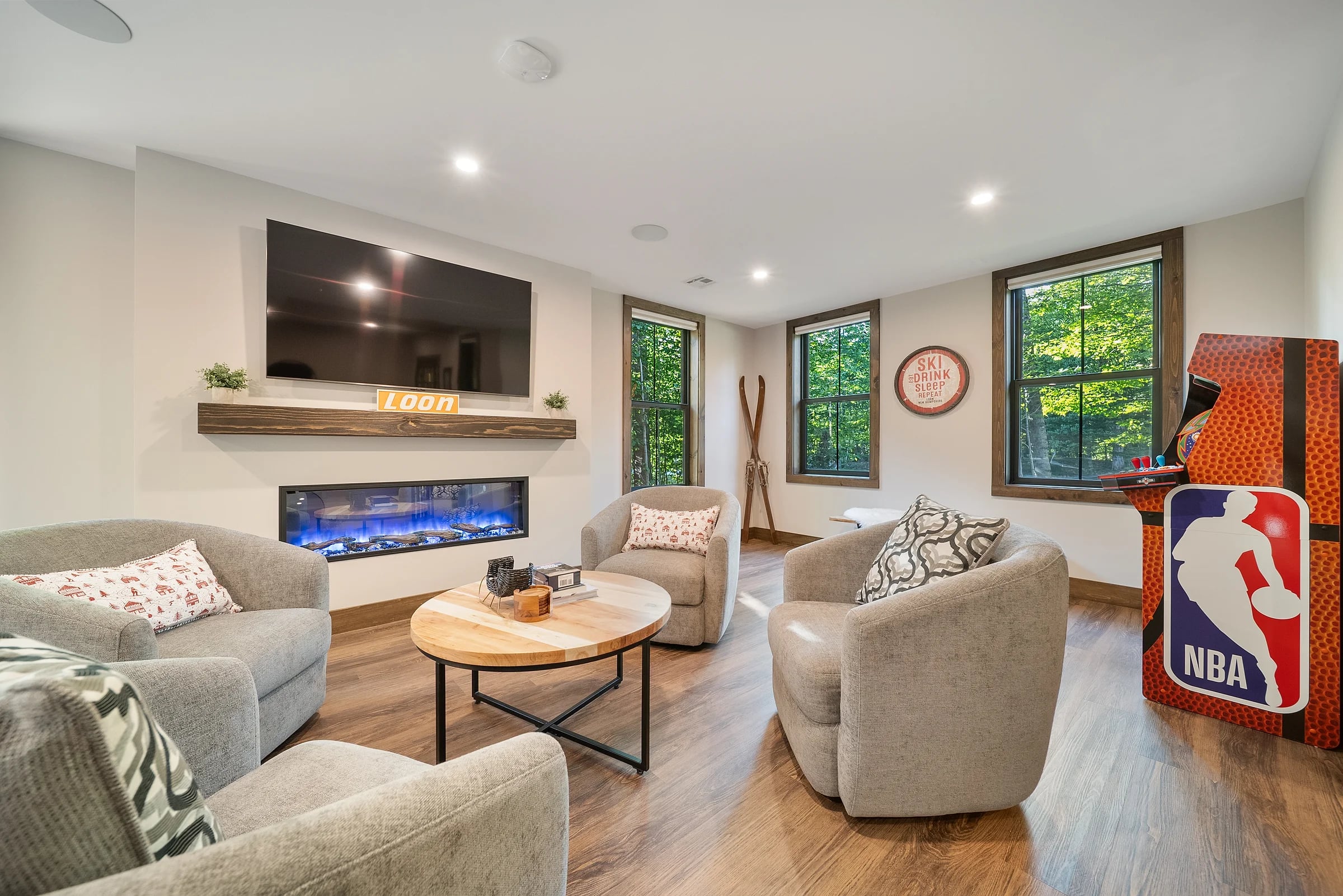 Sitting room with a linear fireplace under a mounted screen, swivel chairs around a round table and forest through the windows
