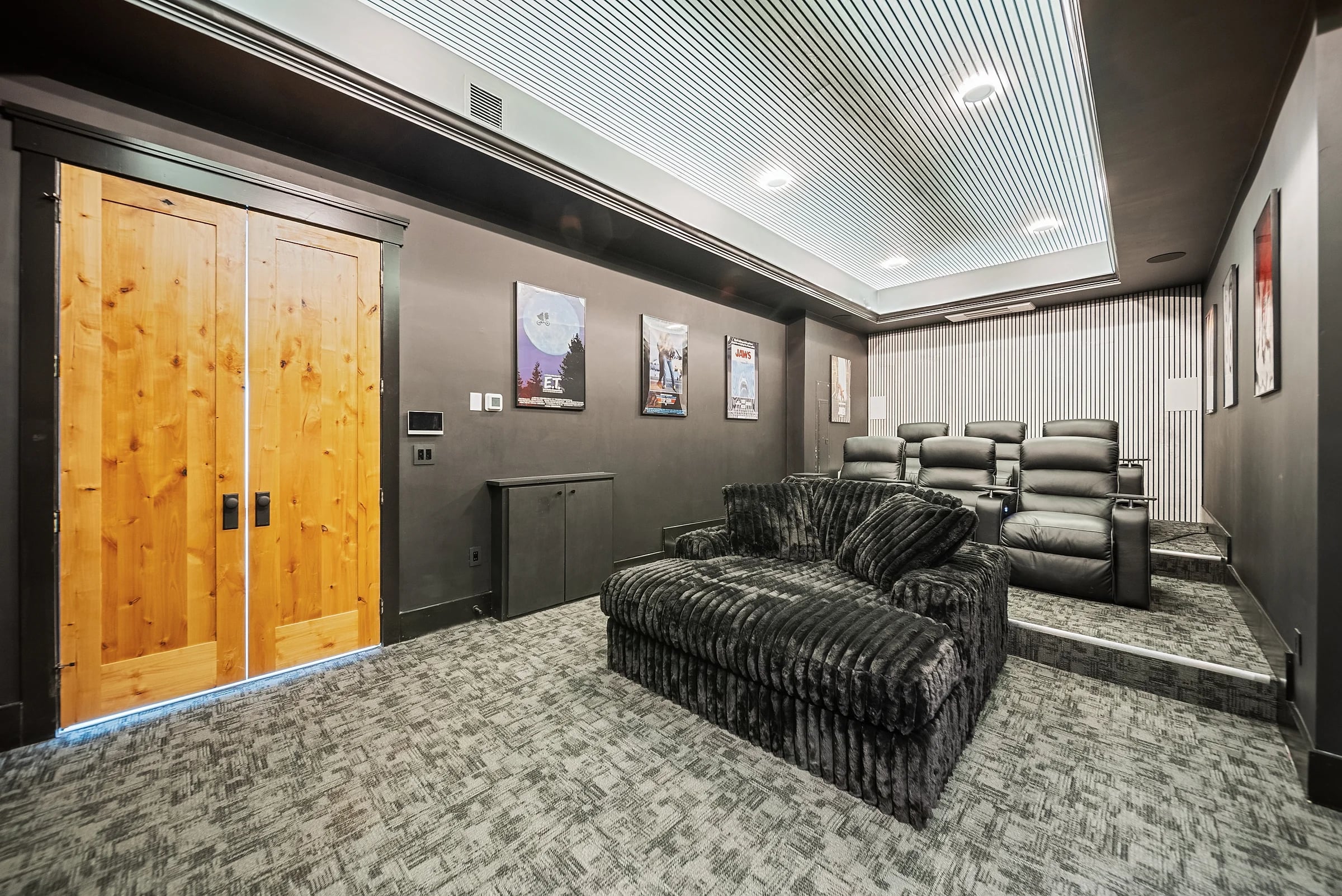 Screening room in charcoal with tiered recliners, a backlit slatted ceiling and knotty-pine double doors at the entry