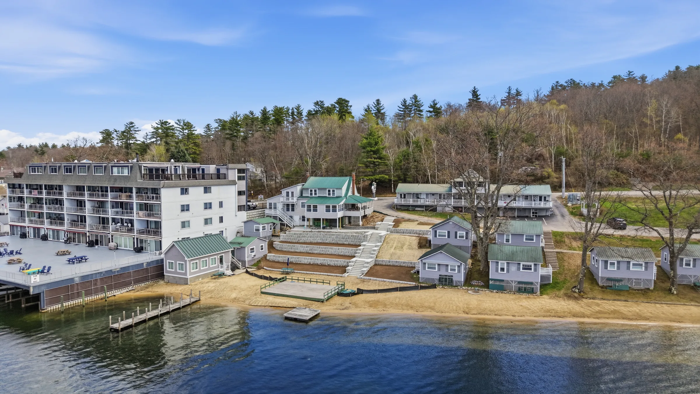 An aerial view of a lakefront resort, a white lodge building with balconies above a sandy beach, guest cottages and docks along the shore