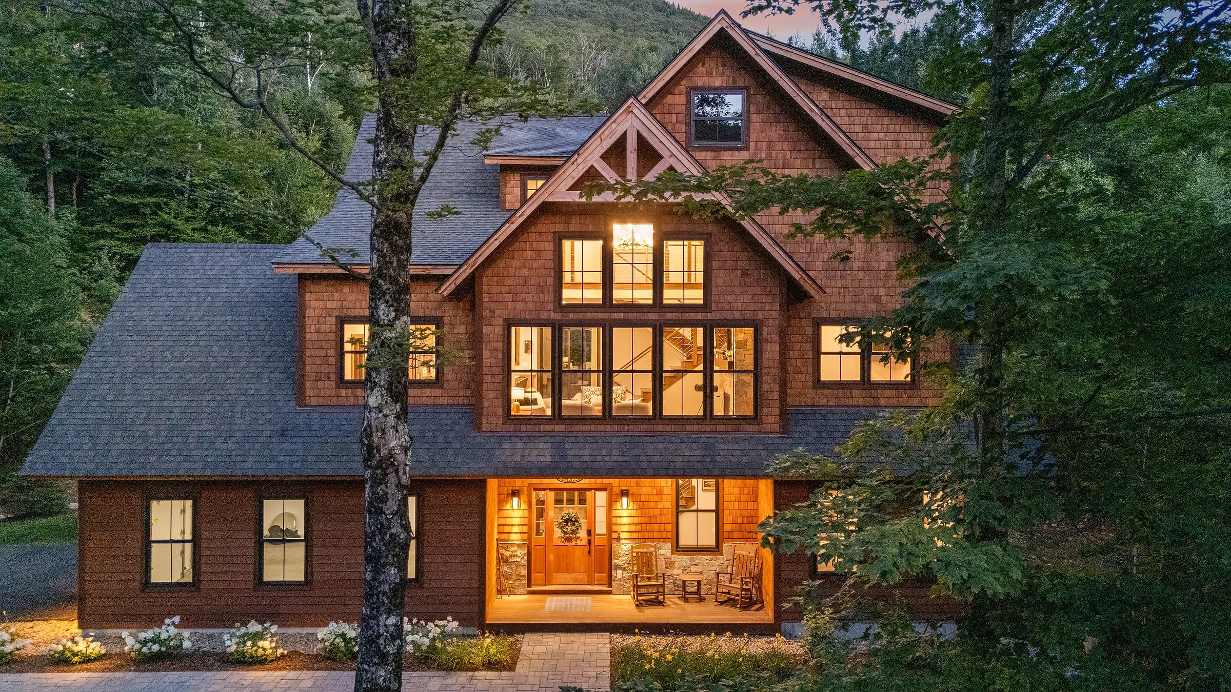 A cedar-shingled lodge lit warm at dusk, gables and a covered entry set against dense forest