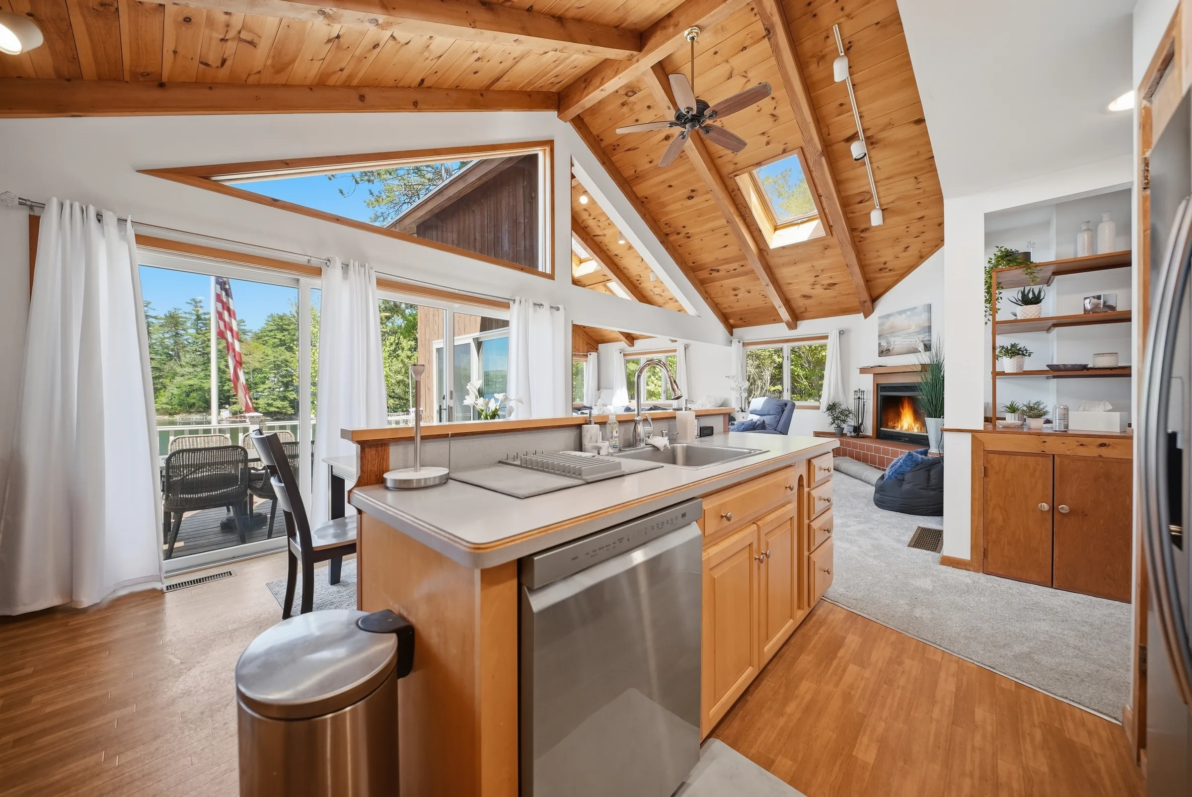An open great room beneath a pine cathedral ceiling, a kitchen island in the foreground and a lit fireplace beyond