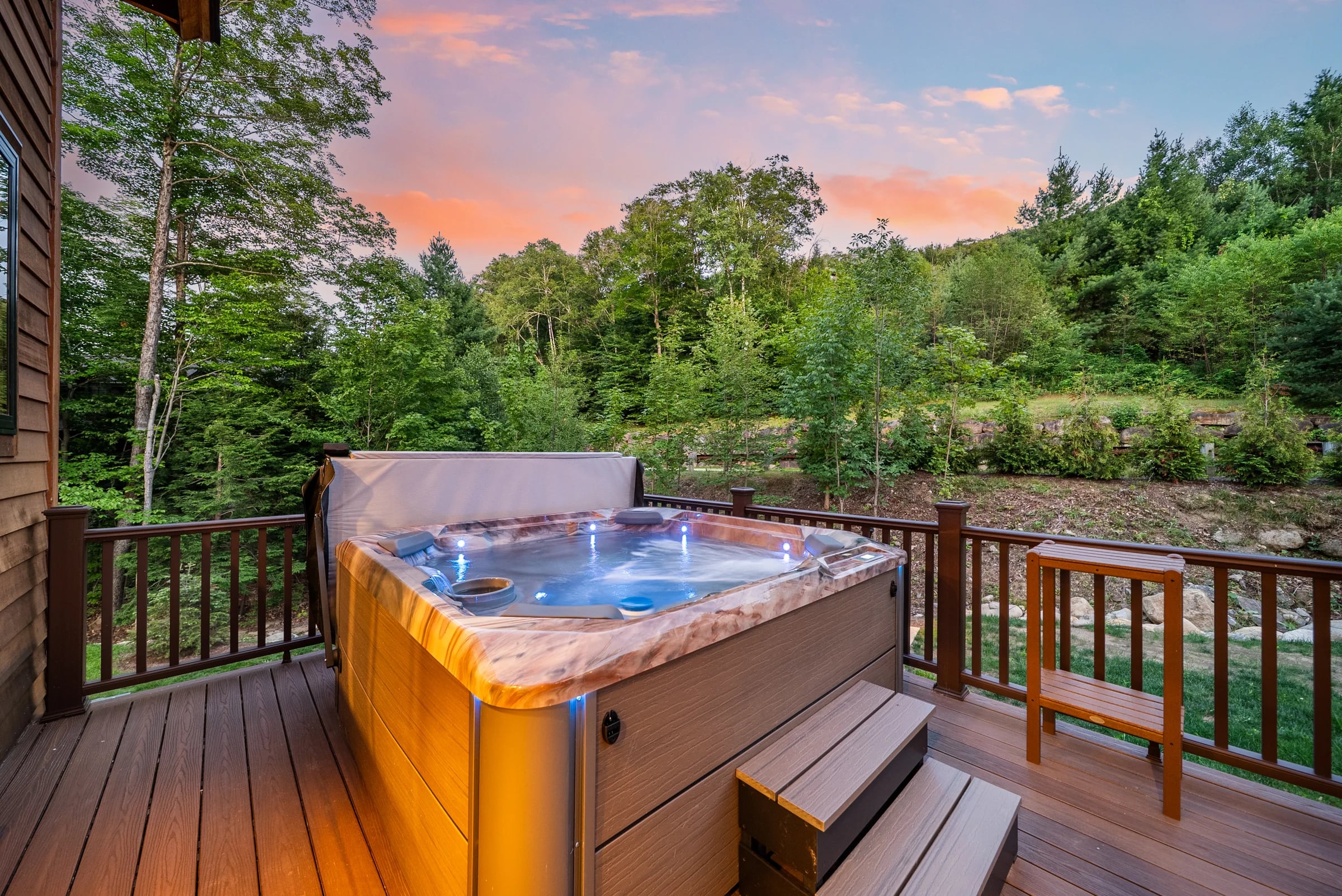A hot tub glowing on a wood deck at dusk, forest and a sunset sky beyond