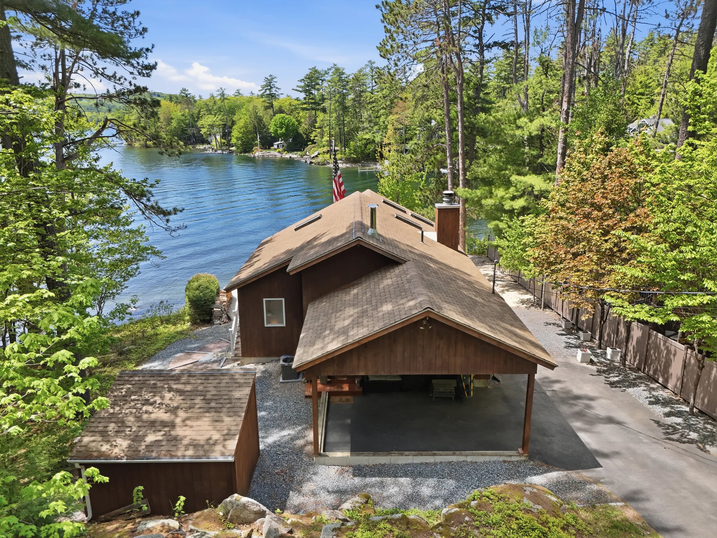 A brown-shingled lakefront cottage and open boathouse on a pine-lined shore, the lake opening out behind