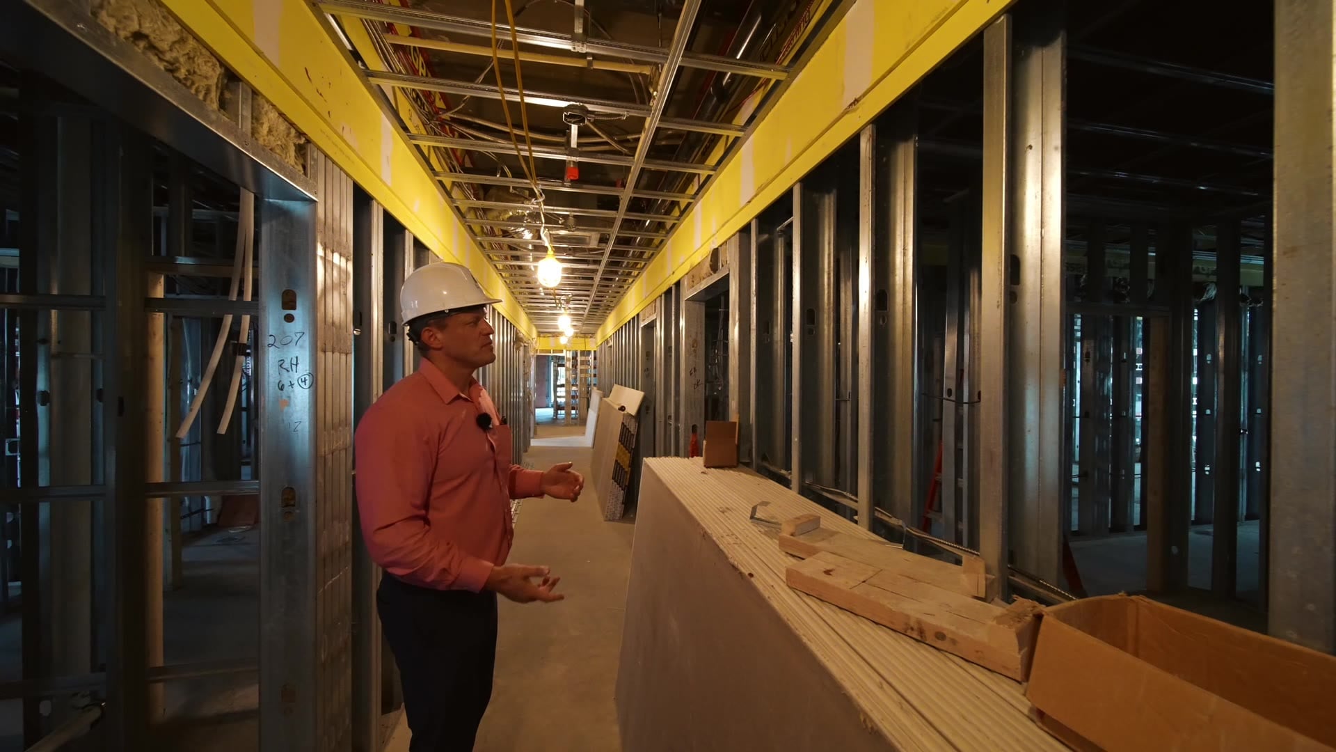 A builder in a hard hat mid-explanation in a light-gauge steel corridor at Grand Central Suites, framing and overhead joists running the length of the floor